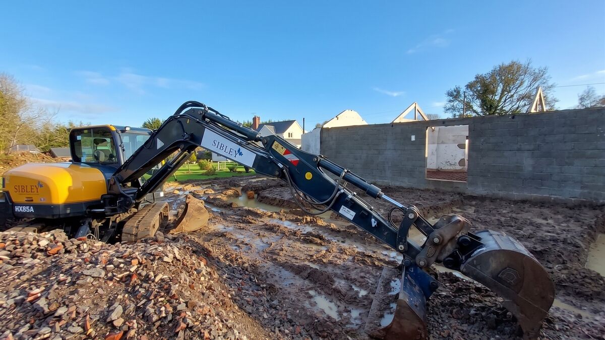 Sibley Contractors excavator digging muddy footings beside a blockwork barn wall in Ashill, Somerset
